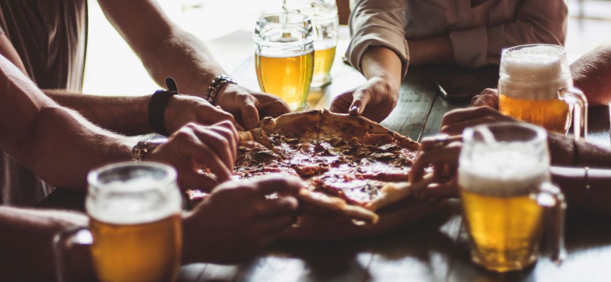 Group of people sharing a pizza at a wooden table with several mugs of beer, highlighting a casual dining and foodie experience.